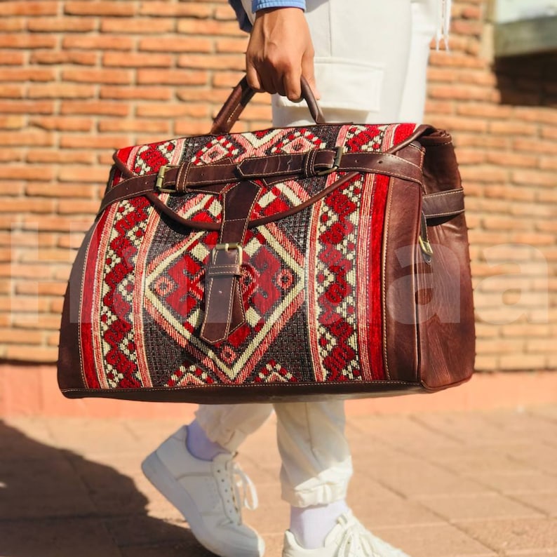 May include: A brown leather travel bag with a red, white and black patterned fabric front. The bag has a large main compartment and a smaller front pocket.