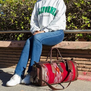 May include: A person wearing blue jeans and a grey sweatshirt with "CALIFORNIA LOS ANGELES" printed on it, sitting on a stone wall with a red and brown leather travel bag with a woven pattern.