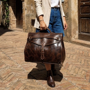 May include: A large, dark brown leather satchel bag featuring a prominent front flap with a vertical buckle strap and a horizontal strap across the top. The bag has a distressed, mottled texture and a sturdy top handle, showcasing a classic design.