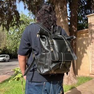 May include: A black leather backpack with adjustable straps and buckles. The backpack has a roll-top closure and a flap with a buckle.