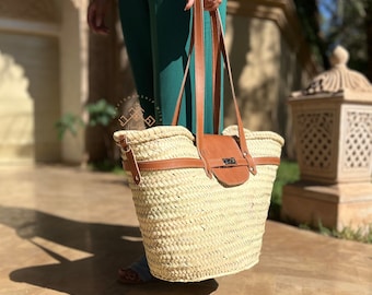 May include: A large, woven straw tote bag with brown leather handles and a brown leather flap closure. The bag has a woven pattern and is a light beige color.
