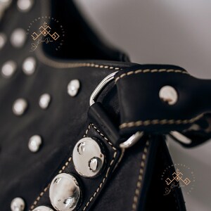 May include: Close-up of a black leather handbag with silver studs and a silver metal buckle. The bag has a brown leather strap with white stitching.