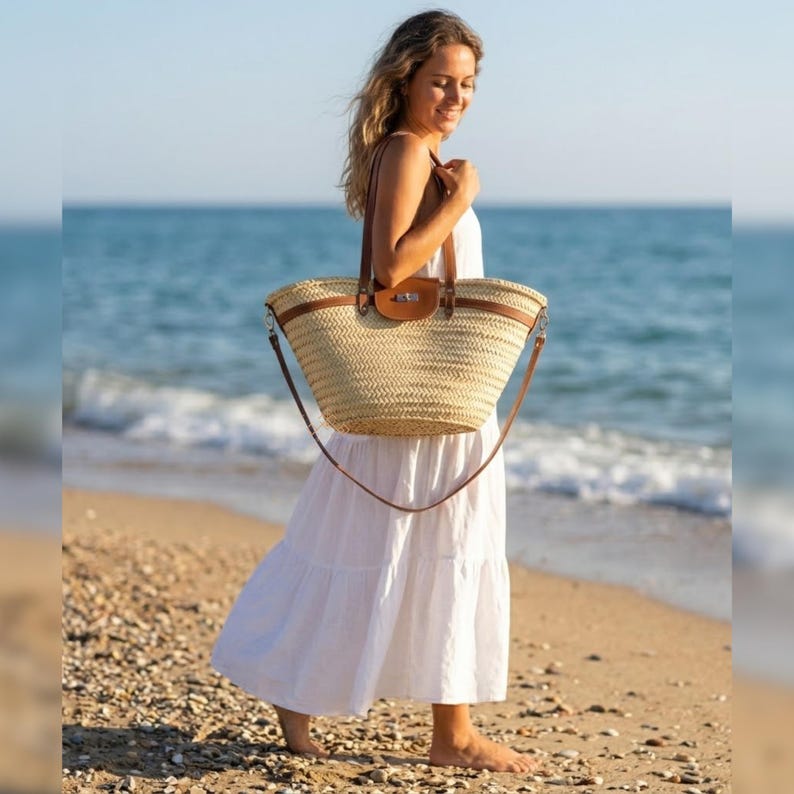 May include: A natural woven straw tote bag with brown leather handles and trim. The bag has a long, adjustable shoulder strap. The bag is being carried by a person wearing a white dress on a beach with the sea in the background.
