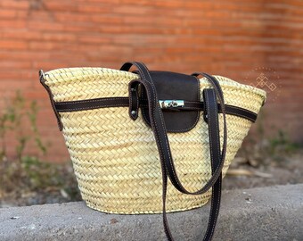 Puede incluir: Un bolso tote de paja tejido con correas de cuero marrón y una solapa de cuero marrón con un cierre de plata. El bolso es de color marrón claro y tiene un patrón tejido.
