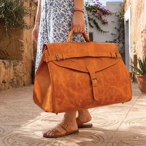 May include: A large, tan leather travel bag featuring a wide front flap with a horizontal strap and a vertical buckle closure. The bag has a structured rectangular shape with visible stitching along the edges and a top handle for carrying.