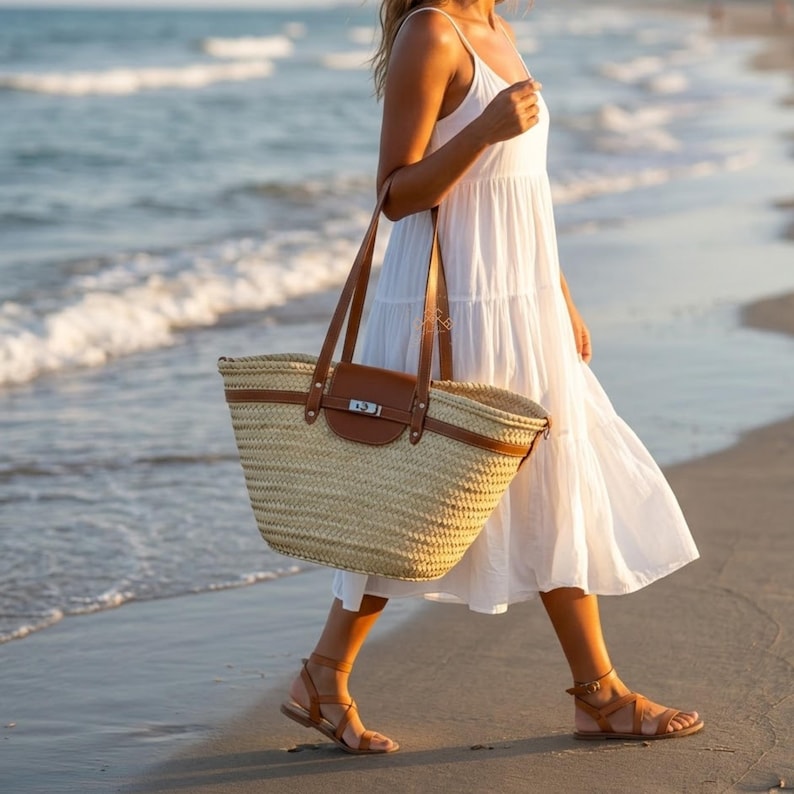 May include: A woven straw tote bag with brown leather details and handles. The bag has a rectangular shape and a brown leather flap closure. The bag is carried by a person wearing a white dress and brown sandals while walking on a beach.