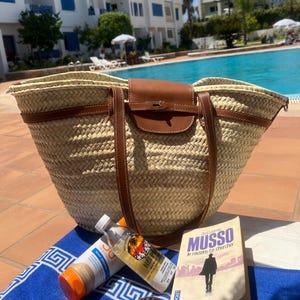 May include: A woven straw tote bag with brown leather details, a flap closure, and shoulder straps. Beside the bag are sunscreen, a bottle of oil, and a book titled "MUSSO". The items are on a blue towel near a swimming pool.