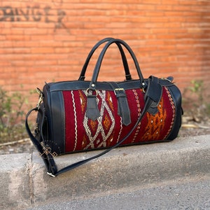 May include: A black leather duffel bag with a red, orange, and white patterned fabric panel. The bag has a top handle and a long adjustable shoulder strap.