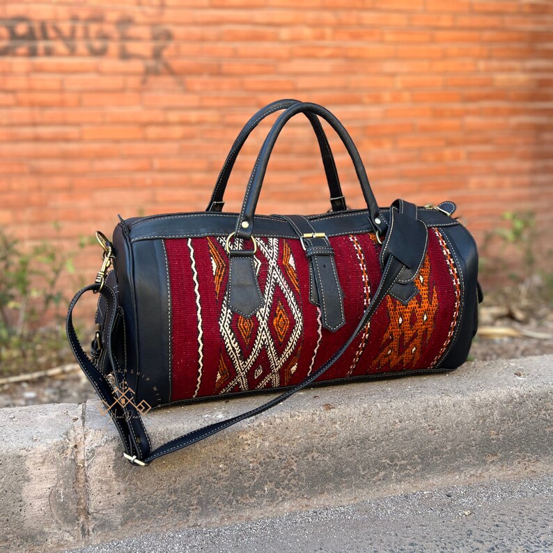 May include: A black leather duffel bag with a red, orange, and white patterned fabric panel. The bag has a top handle and a long adjustable shoulder strap.