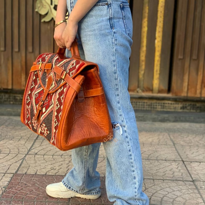 May include: A brown leather satchel with a red, black, and white patterned flap. The bag has a buckle closure and a strap.