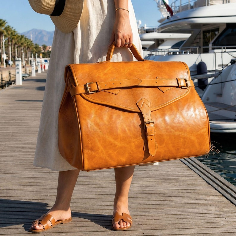 May include: A large, tan leather travel bag featuring a top handle, a wide front flap with a horizontal strap, and a vertical buckle closure. The bag has a smooth, slightly distressed finish and metallic hardware, providing a structured appearance.