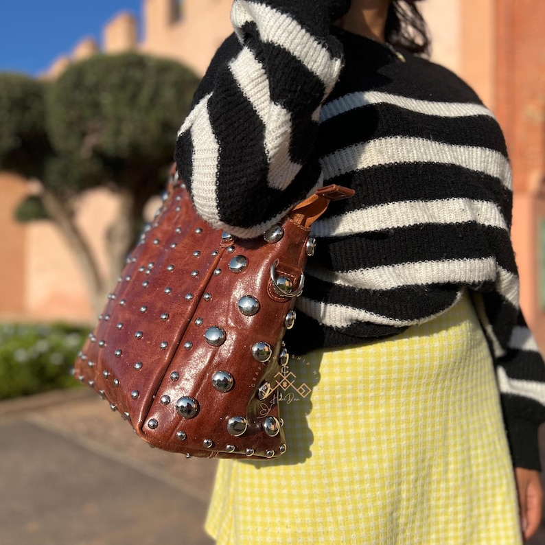 May include: A brown leather tote bag with silver studs. The bag is worn over the shoulder of a person wearing a black and white striped sweater and a yellow gingham skirt.