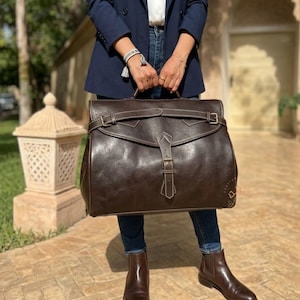 May include: A large dark brown leather satchel bag featuring a wide front flap with a central vertical strap and buckle closure, complemented by a horizontal strap across the top. The bag has visible stitching and metallic hardware accents.
