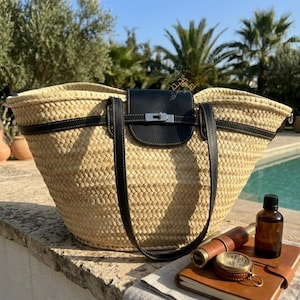 May include: Woven straw tote bag with black leather detailing, including a flap closure and shoulder straps. The bag has a silver-tone clasp. The bag is a stylish accessory, set against a natural outdoor backdrop.