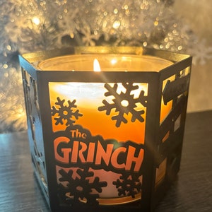 May include: A black metal candle holder with a snowflake design and the words "The Grinch" on the front. The candle is lit and the holder is sitting on a dark surface.