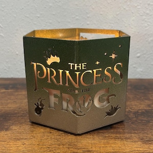 May include: Green and gold candle holder with the text "The Princess and the Frog" cut out. The candle holder has a hexagonal shape and features a crown, stars, and frog silhouettes. The interior is gold, and the candle is visible.