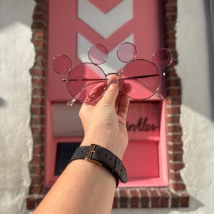 May include: Pink sunglasses with a Mickey Mouse ear design. The round lenses and frames are pink, with three circular accents forming the ears. The sunglasses are held up against a pink background with white chevron shapes.