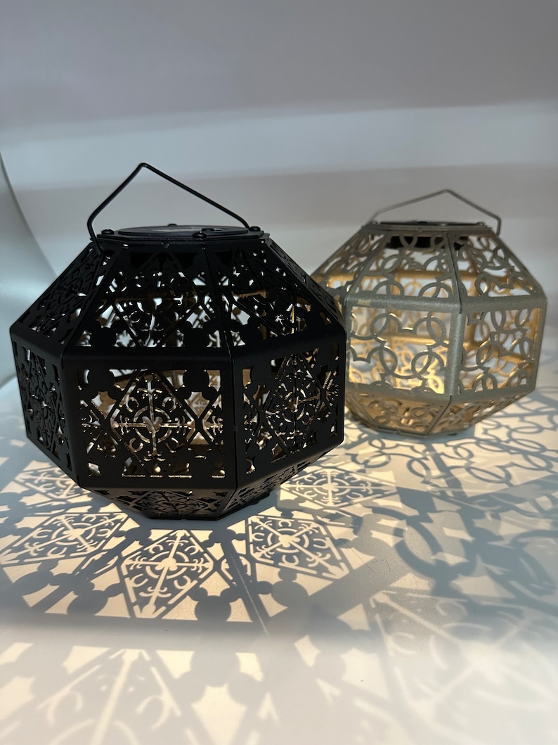 May include: Two decorative metal lanterns with intricate cut-out designs. The lantern on the left is black and the lantern on the right is silver.