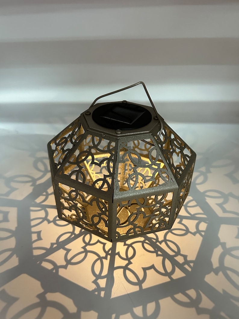 May include: A gold-colored metal lantern with a decorative, intricate design. The lantern has a solar panel on top and casts a patterned shadow on the surface below.
