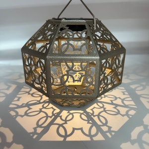 May include: A gold metal lantern with a geometric design and a handle. The lantern casts a shadow on the white surface below.
