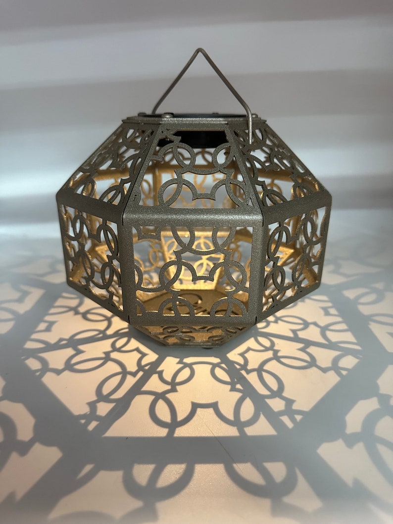 May include: A gold metal lantern with a geometric design and a handle. The lantern casts a shadow on the white surface below.