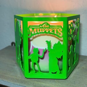 May include: A green laser-cut candle holder featuring The Muppets. The holder has a hexagonal shape with cut-out silhouettes of characters and the show's logo. The candle inside emits a warm glow, creating a cozy ambiance.