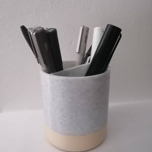 May include: A white ceramic pencil holder with a speckled design and a cream-colored bottom. The holder is filled with various pens and pencils.