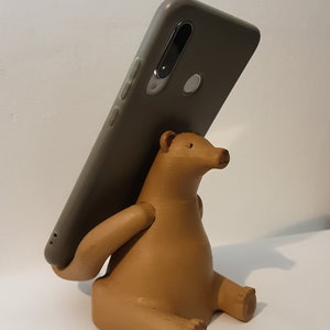 May include: A brown ceramic phone stand shaped like a bear sitting with its arms outstretched, holding a grey smartphone.