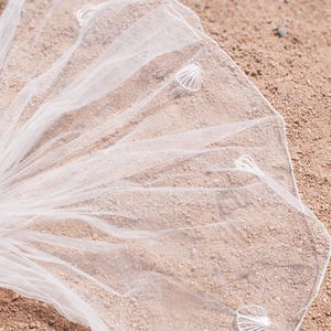 May include: A white bridal veil with seashell accents, resting on a sandy surface. The veil is made of a sheer, delicate fabric and is attached to a clear comb.