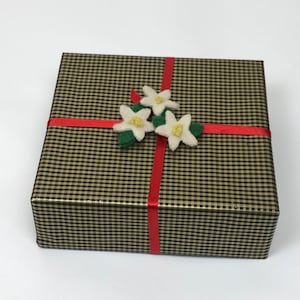 Op de afbeelding: Een zwarte en gouden geruite cadeaudoos met een rood lint en witte viltbloemen.