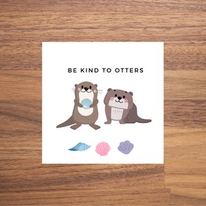 May include: A white card with the text "BE KIND TO OTTERS" in black. Two brown otters are pictured, one holding a seashell. Three seashells are pictured below the otters, one blue, one pink, and one purple.