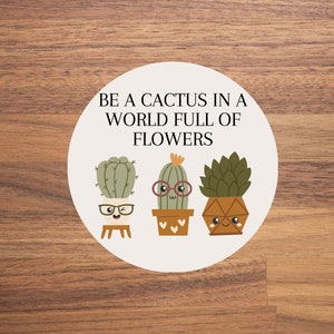 Könnte beinhalten: Ein weißer Kreis mit dem Text "Be a cactus in a world full of flowers" und drei Karikaturkakteen mit Gesichtern und Brillen. Die Kakteen befinden sich in Töpfen und haben unterschiedliche Farben und Designs.