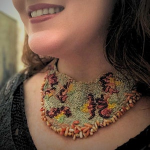 Può includere: Primo piano di una donna che indossa un girocollo ricamato con fiori verdi, rosa e gialli, con una fila di piccole perline rosa chiaro e bianche.