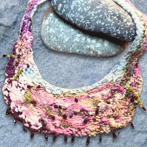 May include: A colourful crocheted necklace with a pink, white and green pattern. The necklace has a fringe of dark red beads and is draped over two grey stones.
