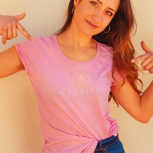 May include: A woman wearing a pink t-shirt with a white logo that says "Creatrix" and a geometric design. She is wearing blue jeans and has her hair pulled back.