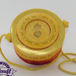 May include: A round, yellow and red decorative box with gold accents and a yellow rope handle. The box has text that reads "I yet outrun the sun. I mark the hours, every one, nor have does not do to". A banner of purple and teal pennants hangs above.