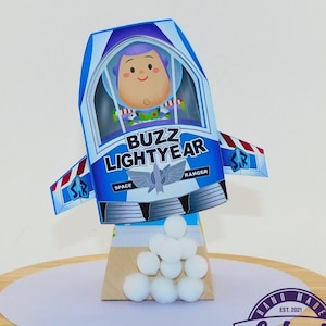 May include: A handmade Buzz Lightyear-themed decoration. The blue and white rocket ship features the character's image and the words "Buzz Lightyear." The decoration is on a wooden base with white pom-poms and a banner of teal and purple pennants.