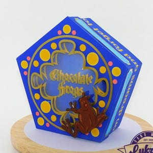 May include: A blue pentagonal box with gold detailing and the words "Chocolate Frogs" is shown. A brown frog figurine is attached to the front. The box sits on a wooden stand, with a banner of purple and teal pennants in the background.