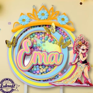 Op de afbeelding: Een meerlagige taarttopper met de naam "Ema" in roze en geel, omgeven door een cirkelvormig frame met kleurrijke confetti. De topper heeft een kroon met blauwe bloemen en gouden vlinders. Een cartoonfiguur van een meisje in een jurk staat rechts.