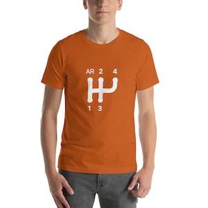 May include: Orange short-sleeved t-shirt with a white graphic of a manual transmission gear shift pattern. The pattern includes the letters "AR" for reverse, numbers 1, 2, 3, and 4, and a stylized gear shift pattern.