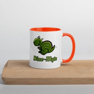 Può includere: Tazza in ceramica bianca con bordo e manico arancioni. La tazza presenta un dinosauro verde dei cartoni animati con il testo "Dino-Myte" in verde e nero.
