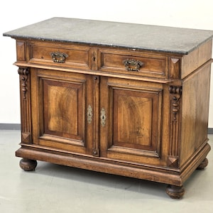 May include: A wooden cabinet with a grey stone top. The cabinet features two drawers and two doors with decorative hardware. The wood has a warm, brown tone, and the cabinet rests on four rounded feet. The style is antique.