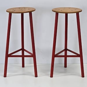 Puede incluir: Dos taburetes altos de bar. Cada taburete tiene un asiento redondo de madera clara con varios agujeros pequeños. Las patas son de color rojo vibrante, con una estructura de soporte triangular cerca de la base. Fondo blanco.