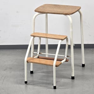 Puede incluir: Un taburete de dos escalones con estructura metálica blanca y escalones de madera. El escalón superior es rectangular y el inferior más pequeño. El taburete tiene patas de goma negra. Fondo blanco.