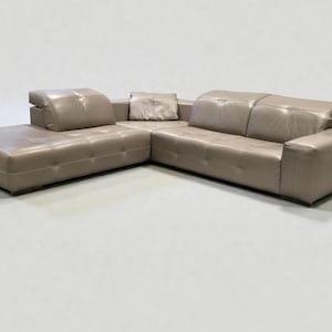 Op de afbeelding: Een taupekleurige hoekbank van leer met een chaise longue aan de linkerkant. De bank heeft een modern design met gecapitonneerde details op de chaise longue en een kussen in de hoek. De bank is neutraal van kleur.