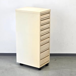 Puede incluir: Un mueble alto de madera clara con múltiples cajones. El mueble es rectangular y se apoya sobre pequeñas ruedas negras. Los cajones son horizontales y están espaciados uniformemente.