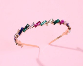 May include: A gold headband with a zig-zag pattern of colorful rectangular rhinestones. The rhinestones are in shades of green, blue, pink, and clear.