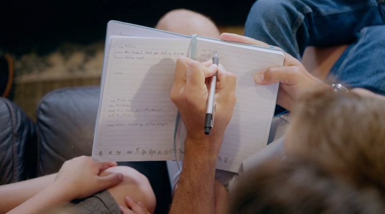 Puede incluir: Una persona escribiendo en un cuaderno de l&iacute;neas con un bol&iacute;grafo plateado. El cuaderno est&aacute; abierto y la persona lo sostiene con su mano izquierda. La mano derecha de la persona sostiene el bol&iacute;grafo y escribe en la p&aacute;gina. El cuaderno tiene una cubierta azul y est&aacute; sentado en el regazo de una persona.