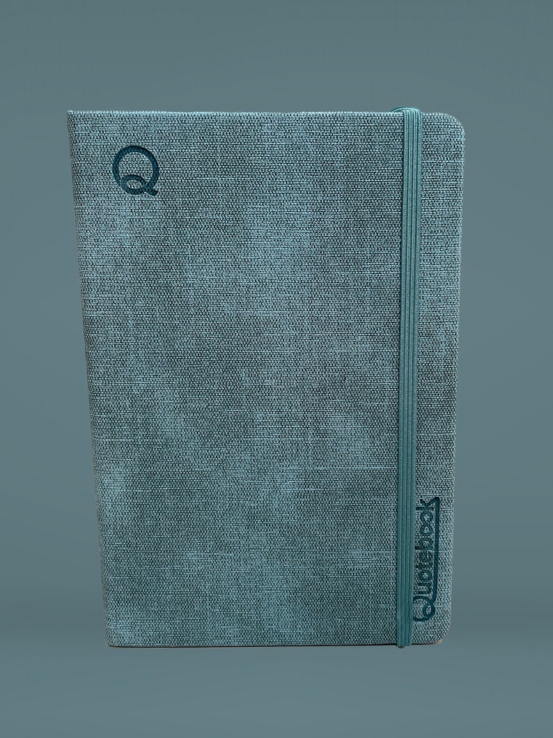 Puede incluir: Un cuaderno gris cubierto de tela con un cierre de banda el&aacute;stica negra y la palabra "Quidebook" impresa en la portada.