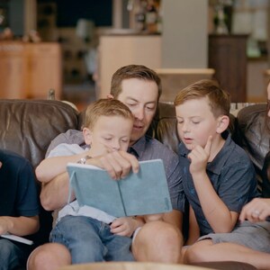 Puede incluir: Una familia de cuatro personas sentada en un sof&aacute; de cuero marr&oacute;n. Un hombre est&aacute; leyendo un libro azul a dos ni&ntilde;os. Una mujer est&aacute; sentada junto a los ni&ntilde;os y sonriendo. El ni&ntilde;o de la izquierda est&aacute; escribiendo en un cuaderno.
